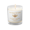 white Christian prayer candle in a clear glass jar with a lit wick decorated with the phrase "Let There Be Light", a gold cross with wings, and the Bible verse Genesis 1:3 printed below.